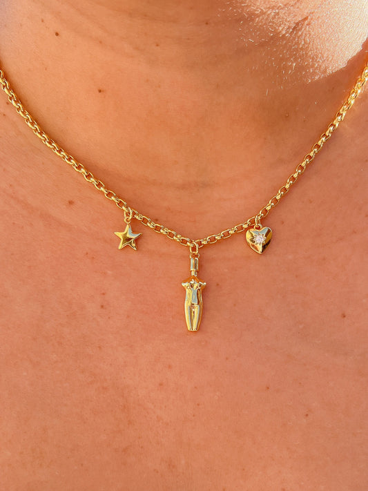 A close-up view of the 14k gold plated Lucia Charm Necklace featuring a center goddess silhouette charm, a star charm, and a cubic zirconia-embedded heart charm worn on a model.