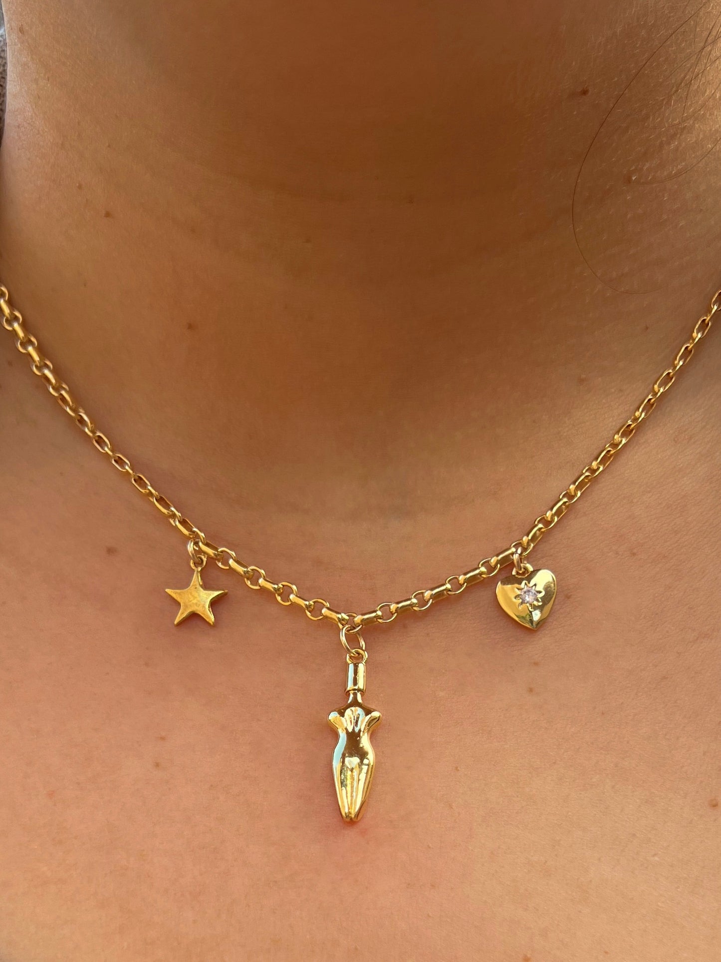 A close-up of model’s neck and chest area wearing the Lucia charm necklace worn. A 3 charm gold-filled necklace with a star (left side), body (center charm), and a cubic zirconia heart charm (right side). Heart charm has a cubic zirconia stone setting in the center of charm.
