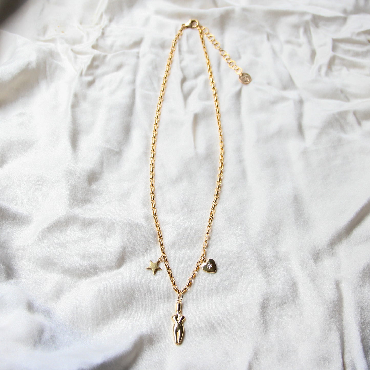 A flat-lay image of a 3 charm gold-filled necklace with an extender chain on a white surface. Necklace can be adjustable from 16 inches to 18 inches. Necklace has a star, body, and a cubic zirconia heart charm.