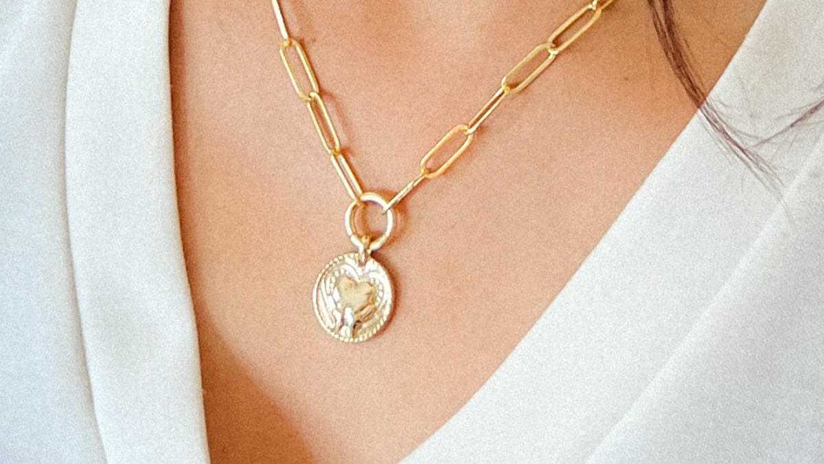 Gold-paperclip necklace with a circle pendant featuring a hand and heart design.