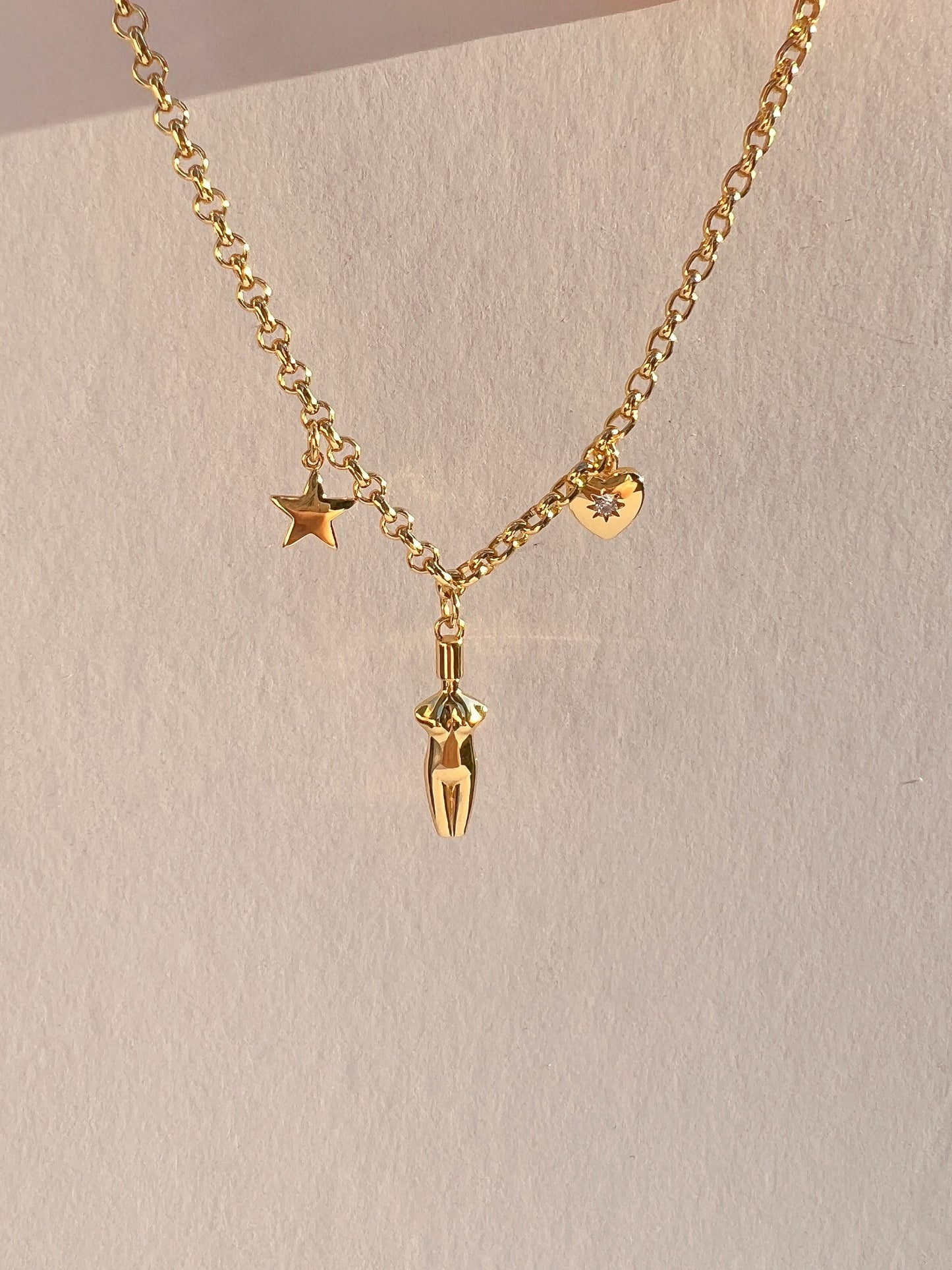Close-up of the Lucia Charm Necklace held against a white background, showcasing the high-shine gold rolo chain and three charms: a polished star, a detailed female torso pendant, and a heart with a cubic zirconia stone setting.