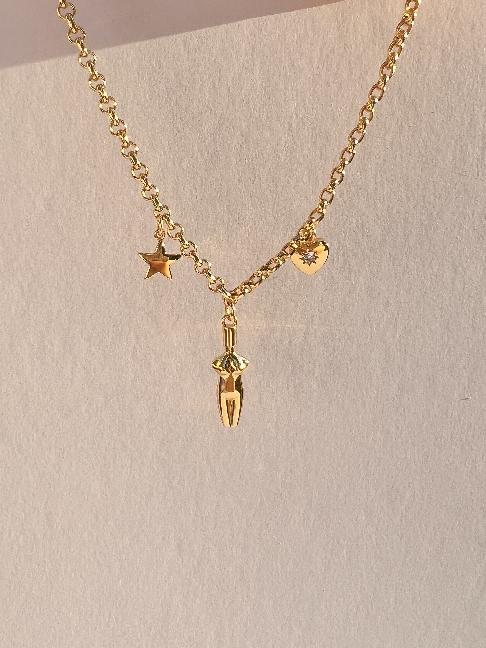 Close-up of the Lucia Charm Necklace held against a white background, showcasing the high-shine gold rolo chain and three charms: a polished star, a detailed female torso pendant, and a heart with a cubic zirconia stone setting.