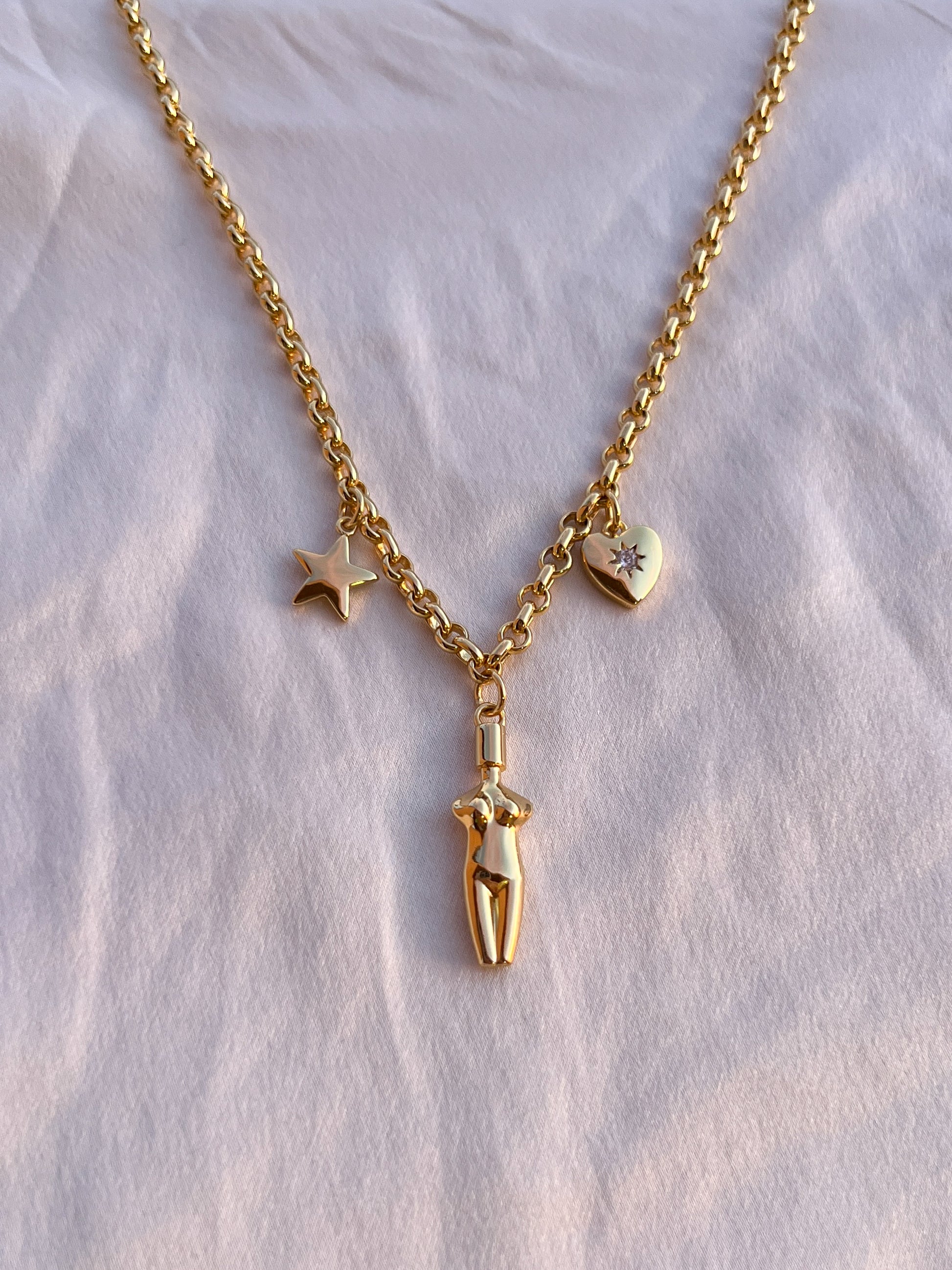 A top-down view of the Lucia Charm Necklace resting on soft white textured fabric, highlighting the intricate chain links and polished charms, star, body, and heart.