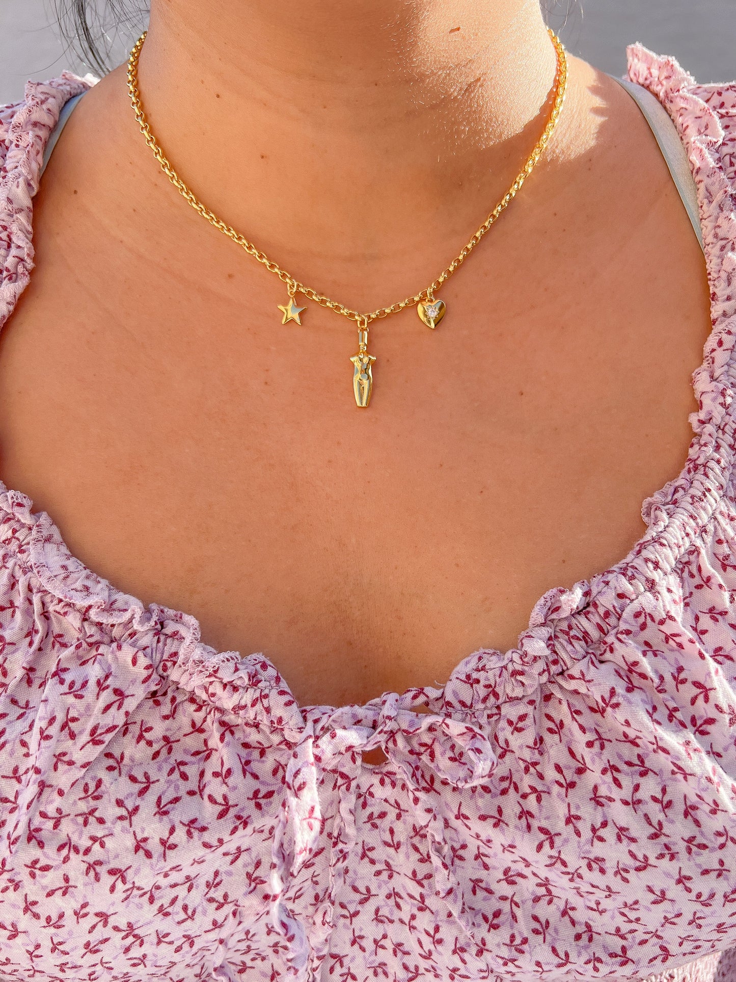 A woman wearing the Lucia Charm Necklace in 14k gold finish, styled with a purple floral flowy dress. The necklace features a gold star, a female body silhouette pendant, and a heart charm with a cubic zirconia stone setting.