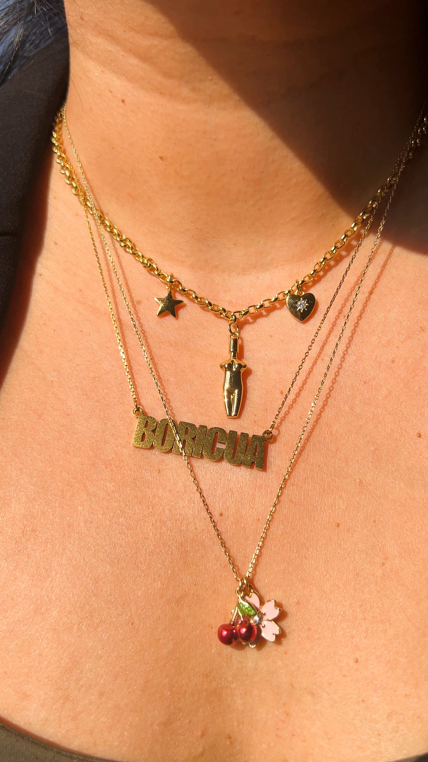 Lifestyle image showing the Lucia Charm Necklace layered with other gold chains, including a "Boricua" nameplate and a cherry charm necklace.