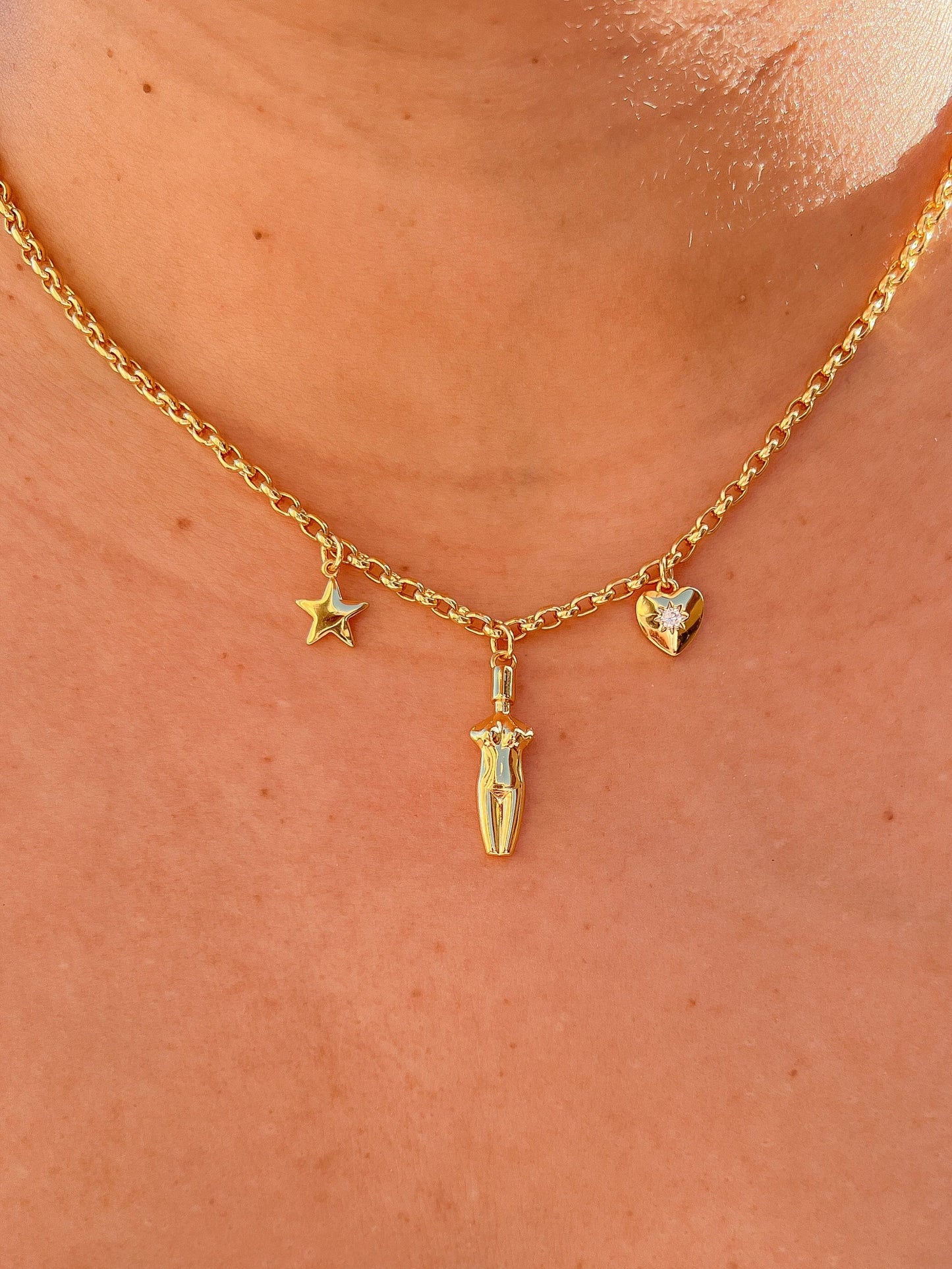 A close-up view of the 14k gold plated Lucia Charm Necklace featuring a center goddess silhouette charm, a star charm, and a cubic zirconia-embedded heart charm worn on a model.