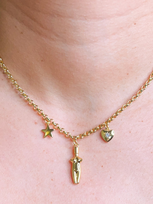 A close-up view of the 14k gold plated Lucia Charm Necklace featuring a center goddess silhouette charm, a star charm, and a cubic zirconia-embedded heart charm worn on a model.