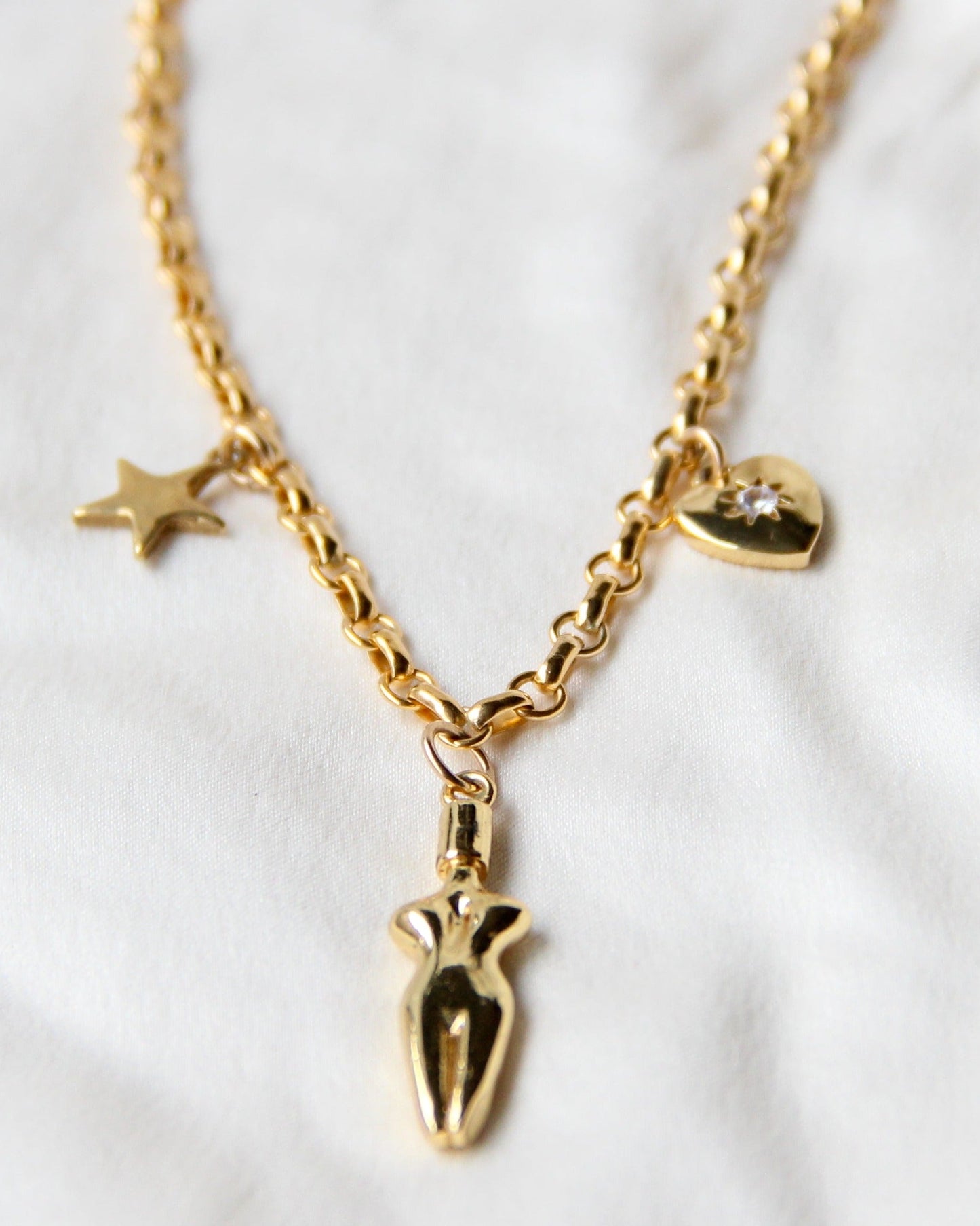 A close-up of the Lucia charm necklace on a silk white fabric background. A 3 charm gold-filled necklace with a star (left side), body (center charm), and a cubic zirconia heart charm (right side). Heart charm has a cubic zirconia stone setting in the center of charm.