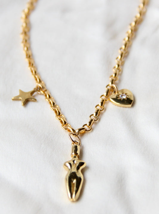 A close-up of the Lucia charm necklace on a silk white fabric background. A 3 charm gold-filled necklace with a star (left side), body (center charm), and a cubic zirconia heart charm (right side). Heart charm has a cubic zirconia stone setting in the center of charm.