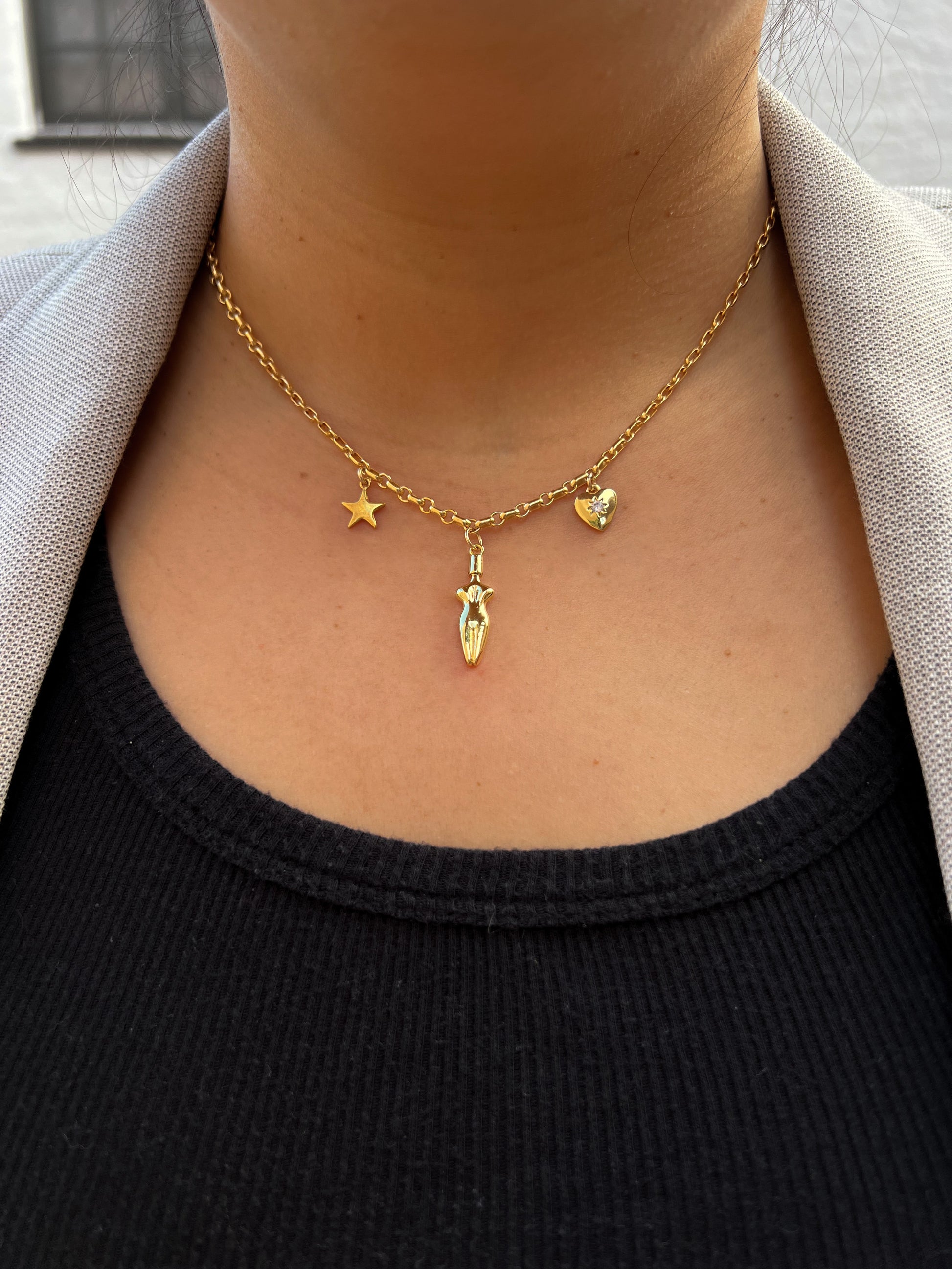 A close-up of model’s neck and chest area wearing a black tank top with a beige/gray blazer and the Lucia charm necklace. A 3 charm gold-filled necklace with a star (left side), body (center charm), and a cubic zirconia heart charm (right side). Heart charm has a cubic zirconia stone setting in the center of charm.
