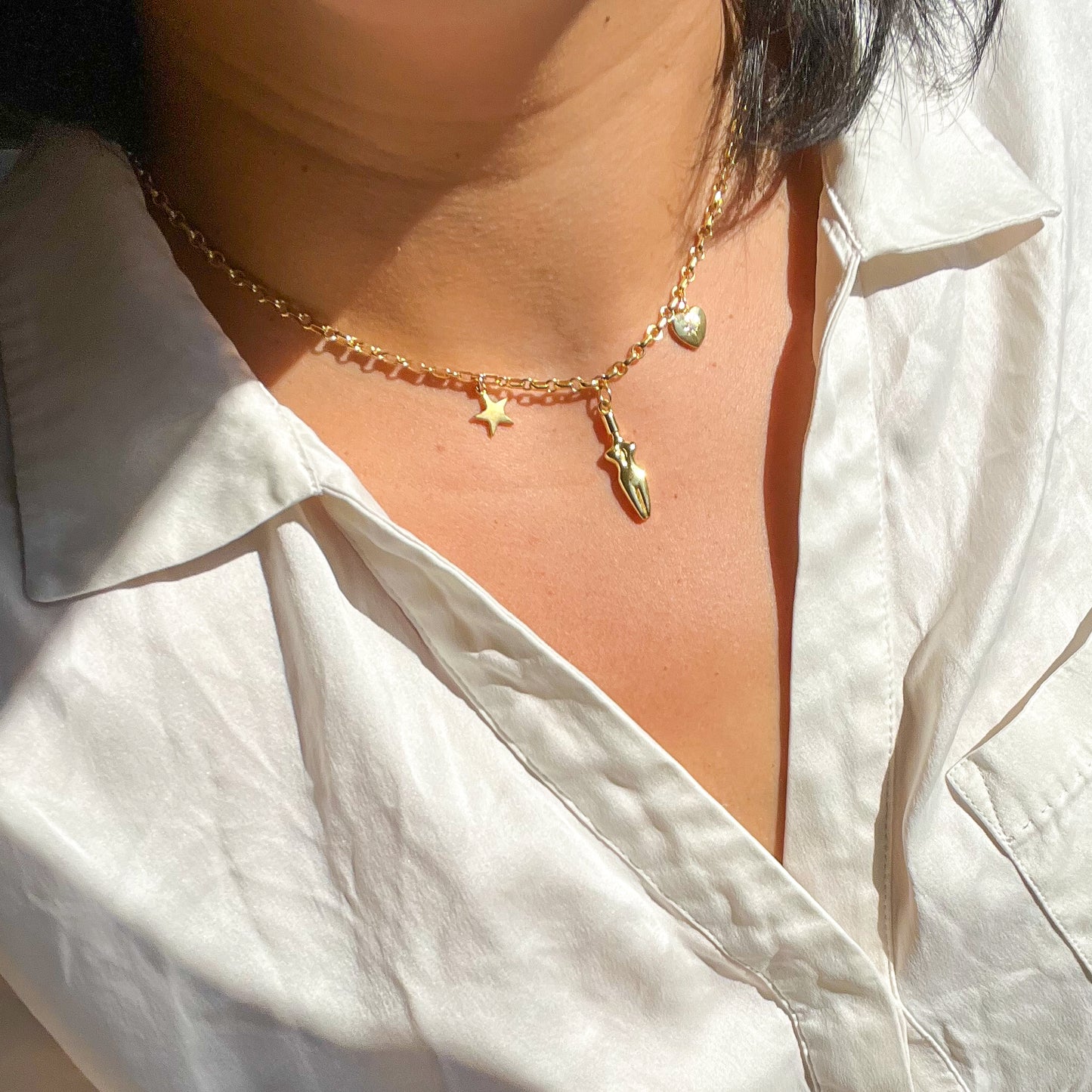 A close-up of model’s neck and chest area wearing a white silk button up shirt and the Lucia charm necklace. A 3 charm gold-filled necklace with a star (left side), body (center charm), and a cubic zirconia heart charm (right side). Heart charm has a cubic zirconia stone setting in the center of charm.