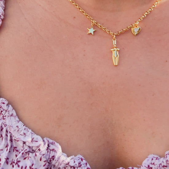 A video of a close-up of a woman wearing the Lucia Charm Necklace in 14k gold finish, styled with a purple floral flowy dress. The necklace features a gold star, a female body silhouette pendant, and a heart charm with a cubic zirconia stone setting.