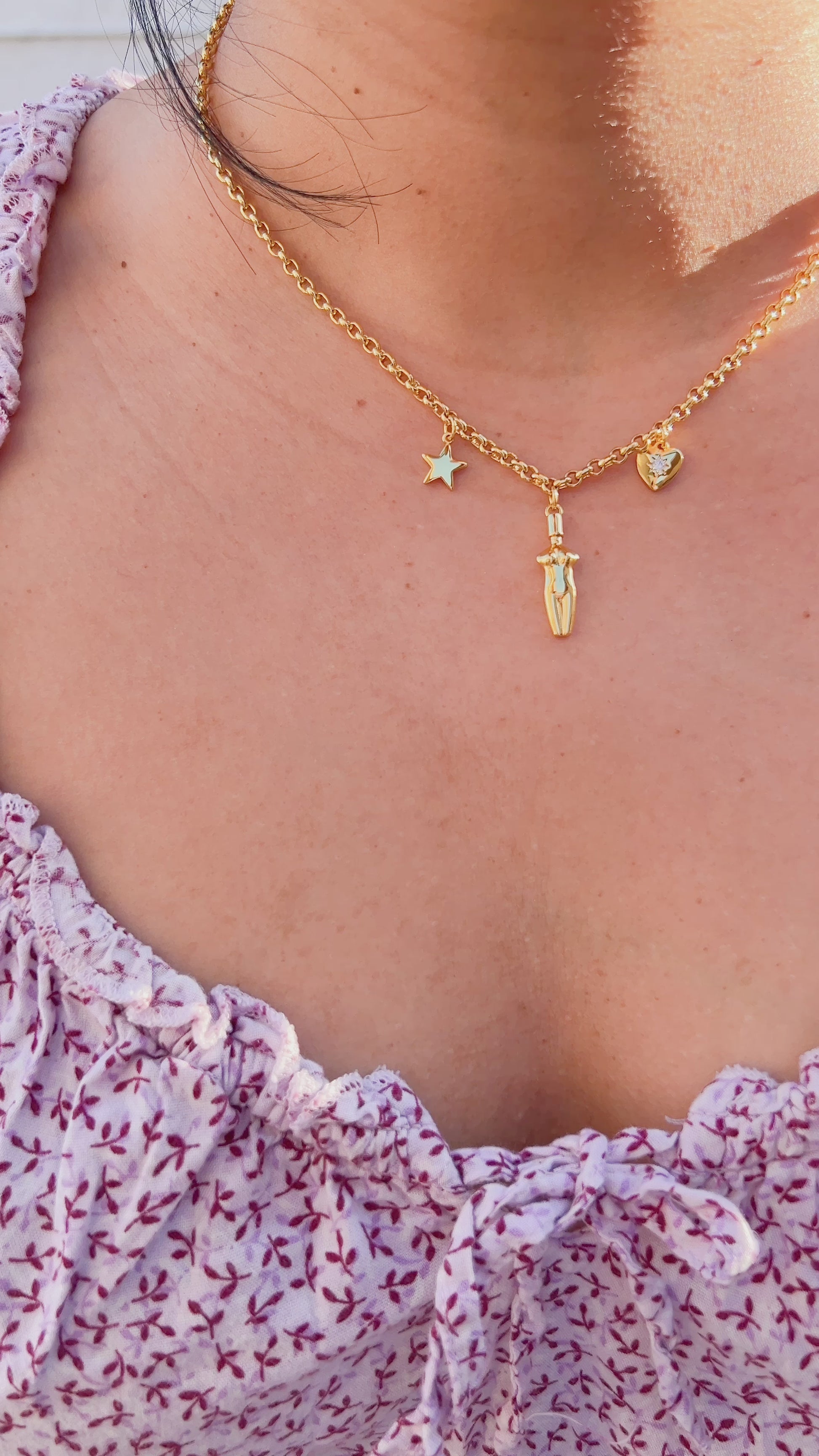 A video of a close-up of a woman wearing the Lucia Charm Necklace in 14k gold finish, styled with a purple floral flowy dress. The necklace features a gold star, a female body silhouette pendant, and a heart charm with a cubic zirconia stone setting.