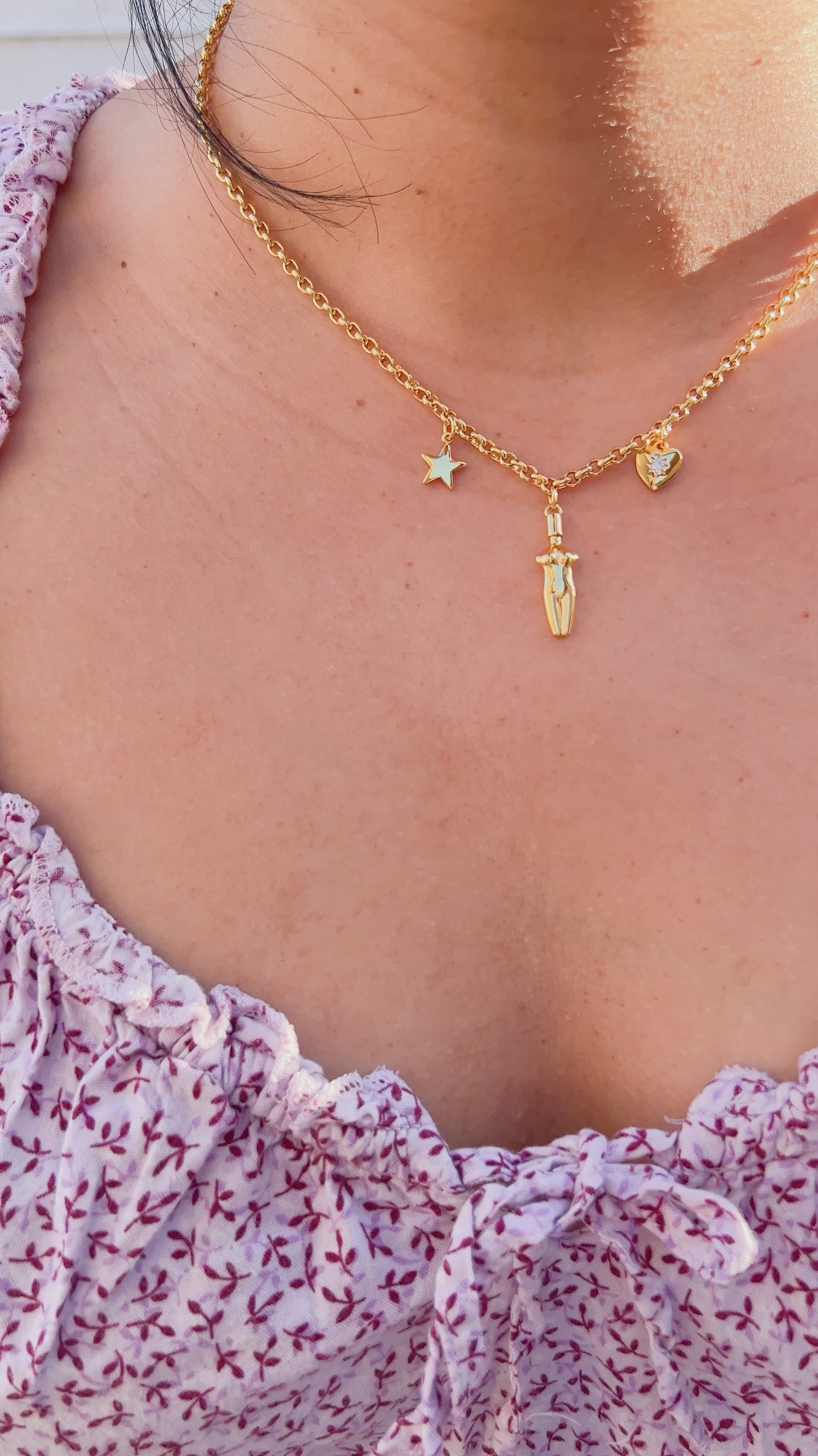 A video of a close-up of a woman wearing the Lucia Charm Necklace in 14k gold finish, styled with a purple floral flowy dress. The necklace features a gold star, a female body silhouette pendant, and a heart charm with a cubic zirconia stone setting.