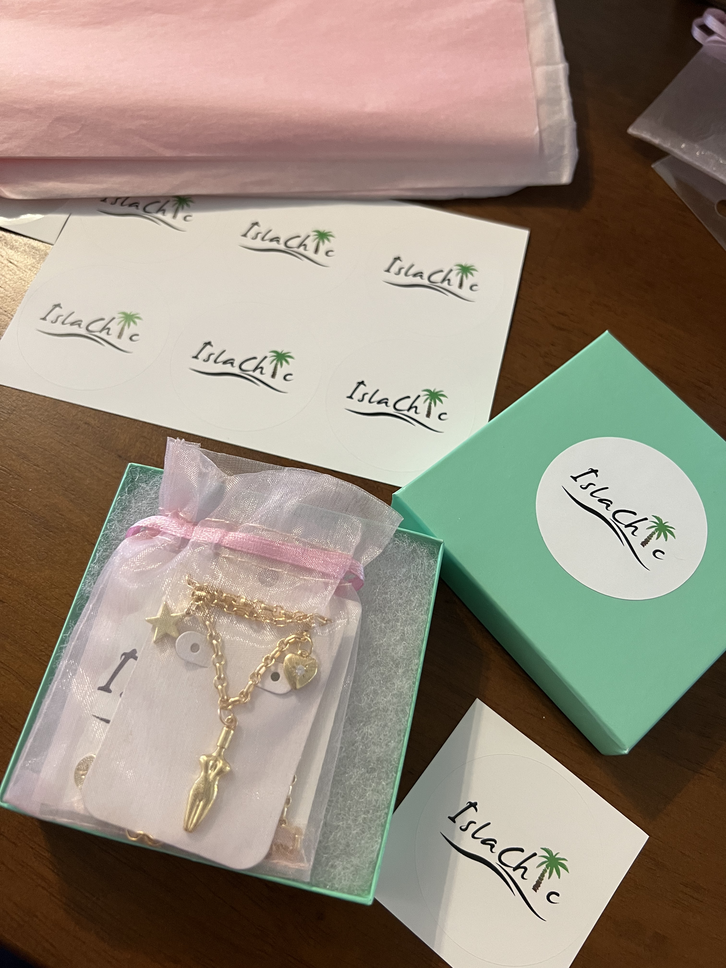 A bird eye view image of our islachic jewelry box/packaging with a 3 charm gold-filled necklace. A teal colored square jewelry box with a circular white sticker with our IslaChic logo. Sitting inside the jewelry box is a pink mesh pouch with our 3 charm gold-filled necklace. Light pink tissue paper and sheet of Islachic logo stickers. 