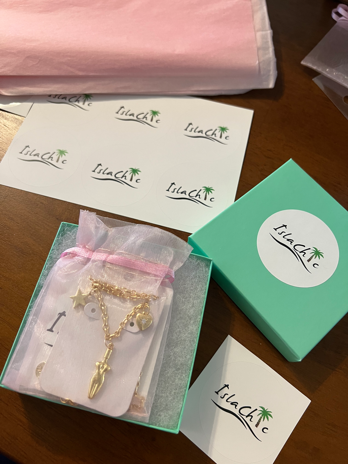 A bird eye view image of our islachic jewelry box/packaging with a 3 charm gold-filled necklace. A teal colored square jewelry box with a circular white sticker with our IslaChic logo. Sitting inside the jewelry box is a pink mesh pouch with our 3 charm gold-filled necklace. Light pink tissue paper and sheet of Islachic logo stickers. 