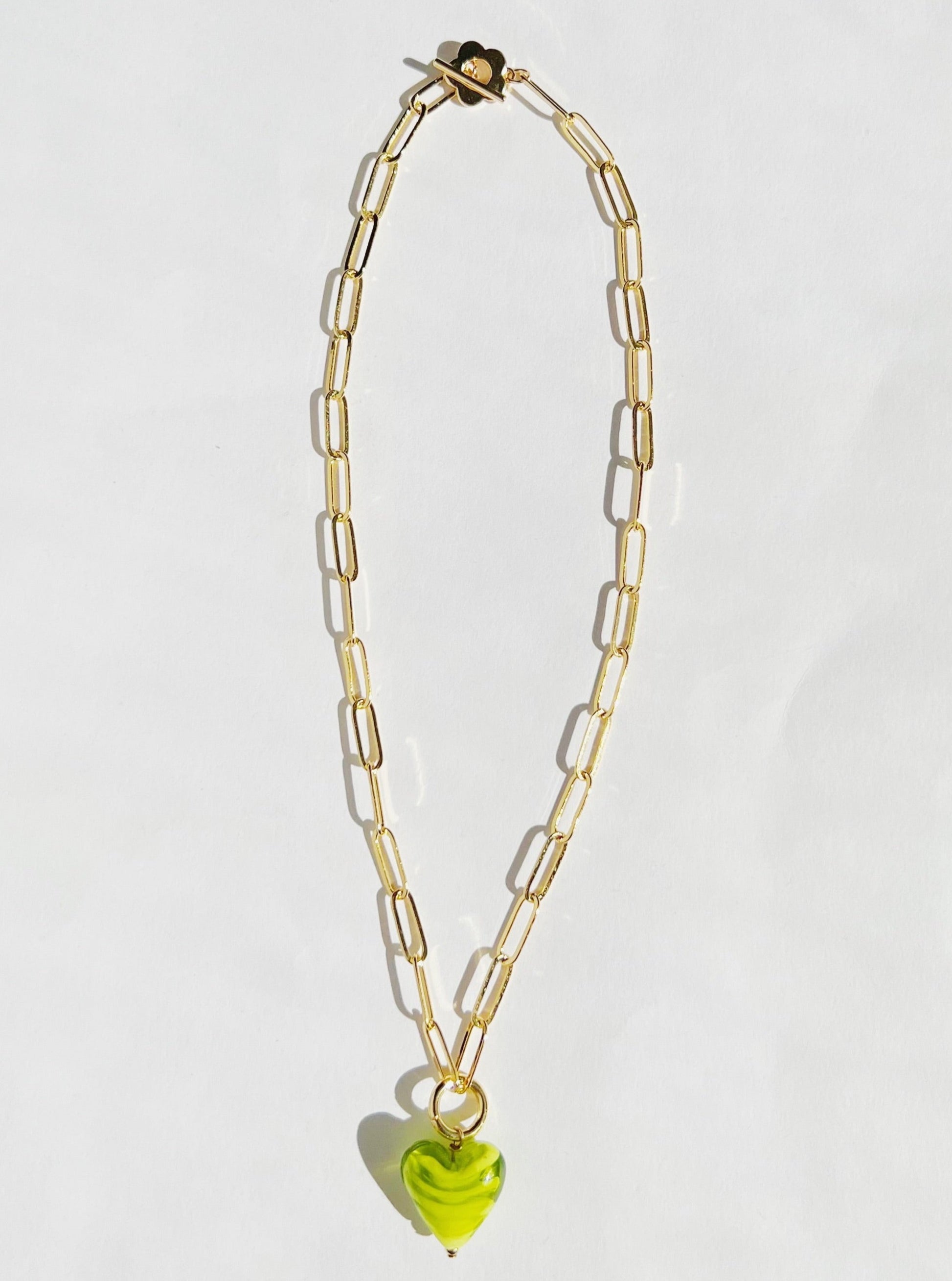 A flat lay picture of a green heart gold paper clip statement necklace on a white background. The statement necklace consist of a glass bead with a green and white spiral color shaped into a heart on a gold paper clip chain with a flower closure.