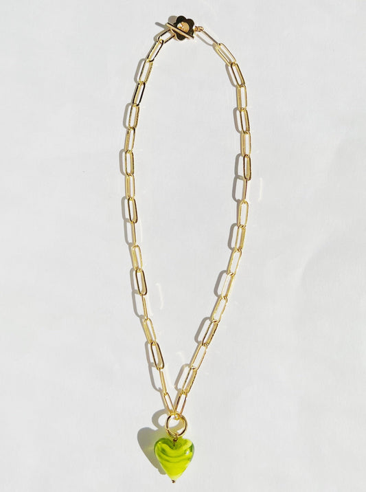 A flat lay picture of a green heart gold paper clip statement necklace on a white background. The statement necklace consist of a glass bead with a green and white spiral color shaped into a heart on a gold paper clip chain with a flower closure.